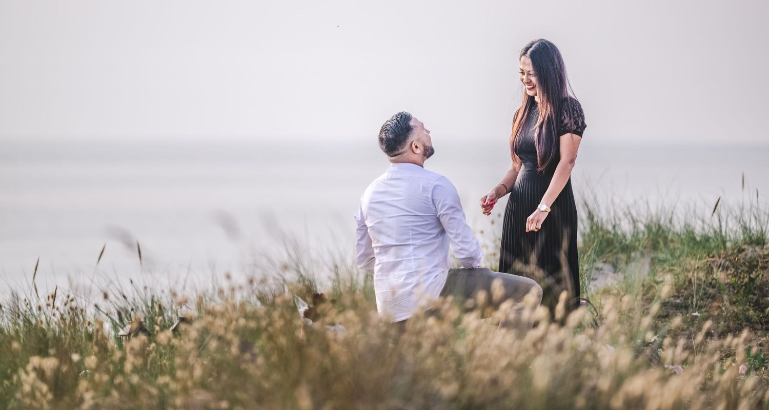 Man proposing on one knee to woman on a scenic coastal clifftop with misty ocean views.