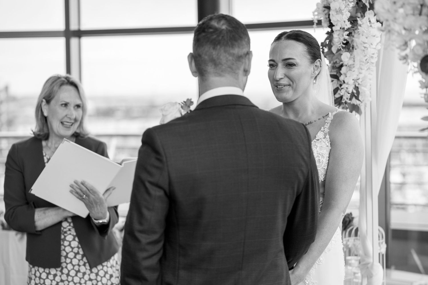 Black and white image of bride smiling at groom during wedding ceremony with officiant beside them.