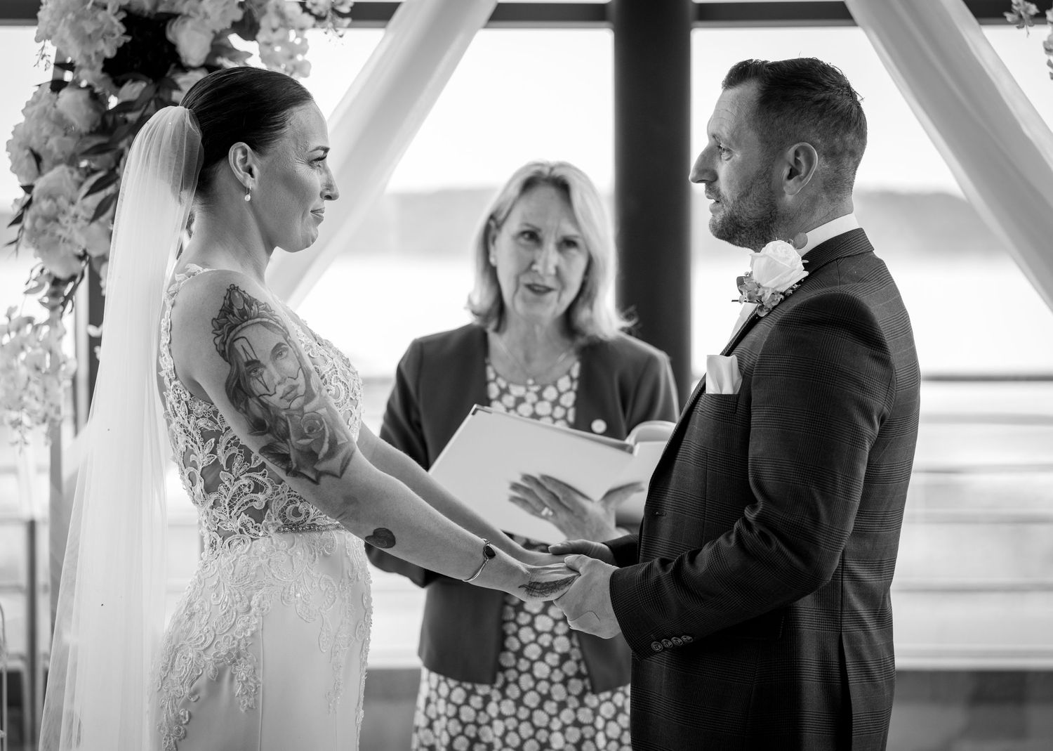 Black and white photo of tattooed bride and groom holding hands before smiling female officiant.