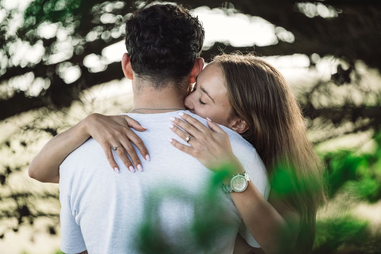 Couple embracing outdoors, woman showing engagement ring while hugging man under trees.