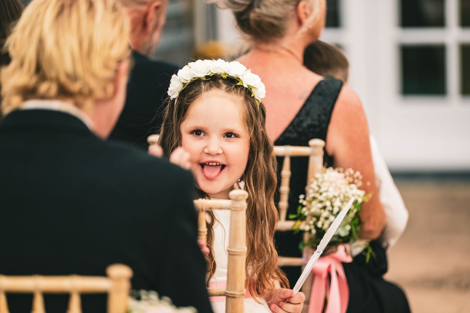 A cheerful flower girl wearing a white floral crown sticks out her tongue playfully during a wedding ceremony.