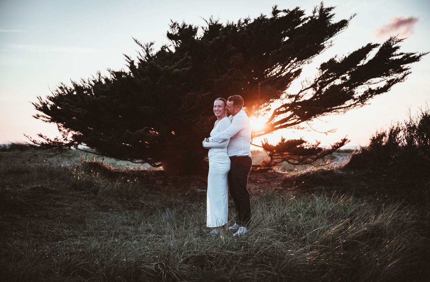 Romantic couple embracing at golden sunset beside a windswept tree on a coastal grassy landscape.