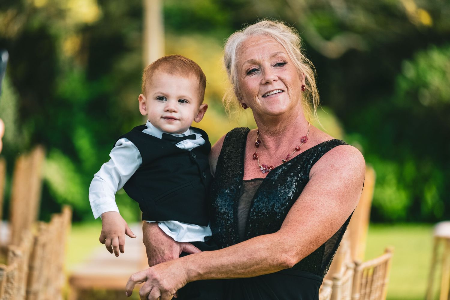 Blonde grandmother in black dress holds baby boy in vest at outdoor wedding reception.