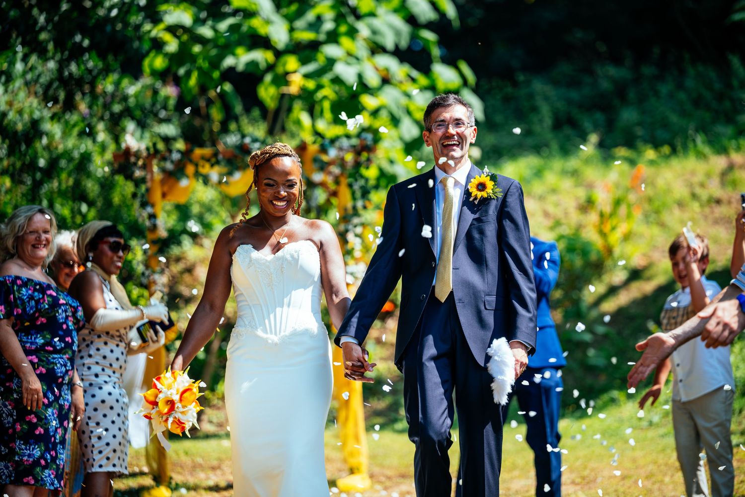 A newlywed couple walks hand-in-hand through a shower of confetti thrown by smiling guests at an outdoor wedding.