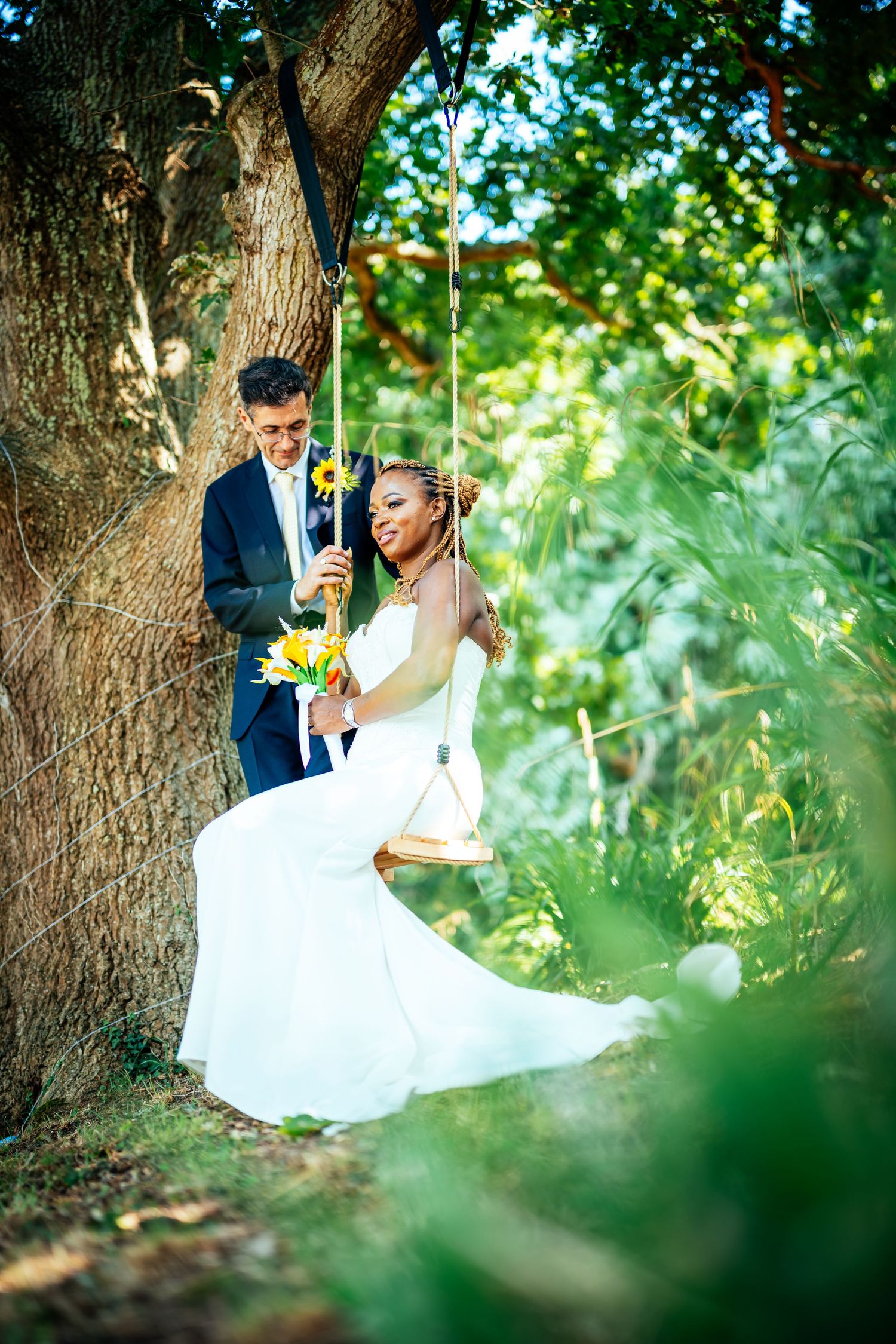 A bride in a white gown sits on a tree swing holding sunflowers while her groom stands beside her in a lush garden.