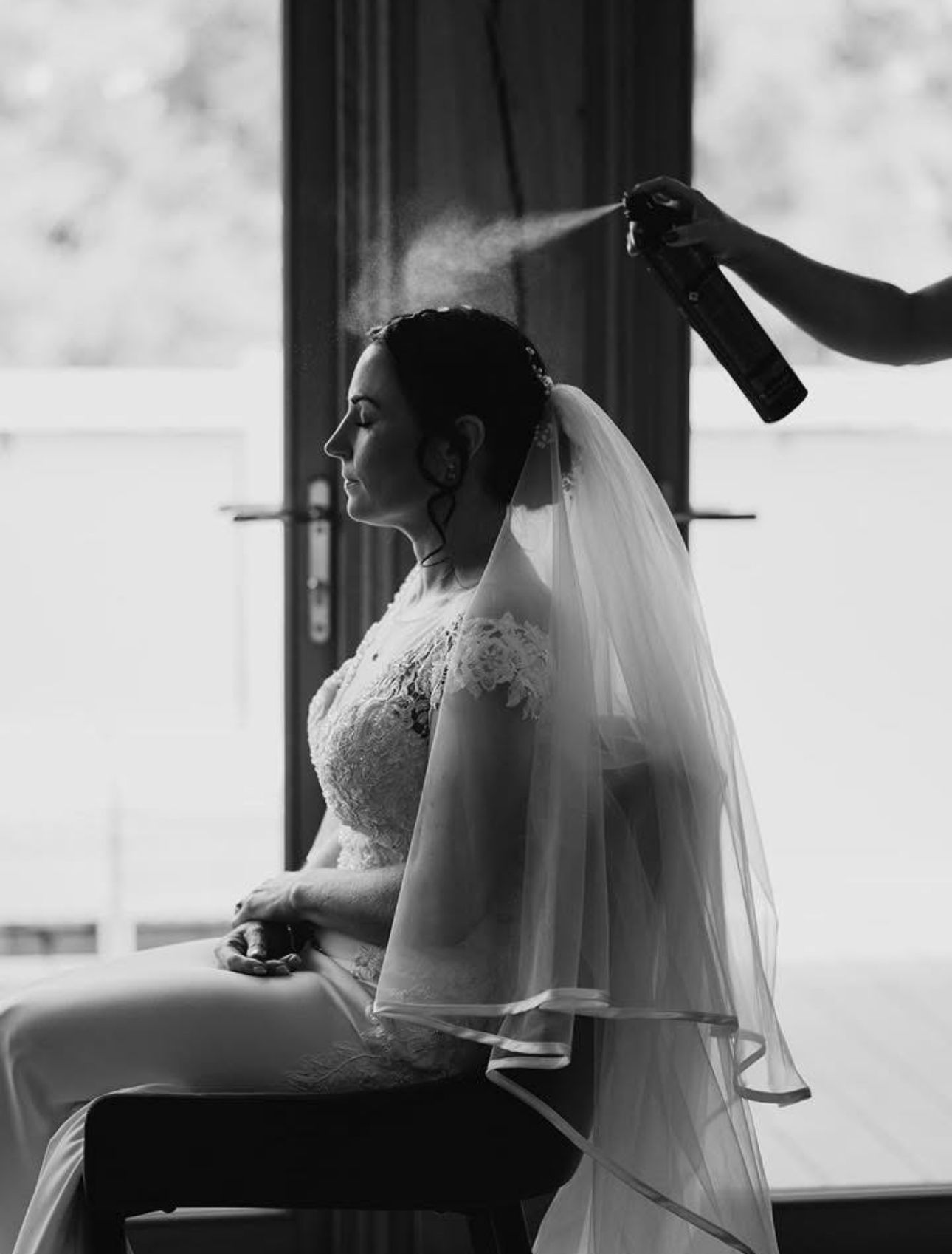 Black and white portrait of bride in veil getting hair styled with spray before wedding.