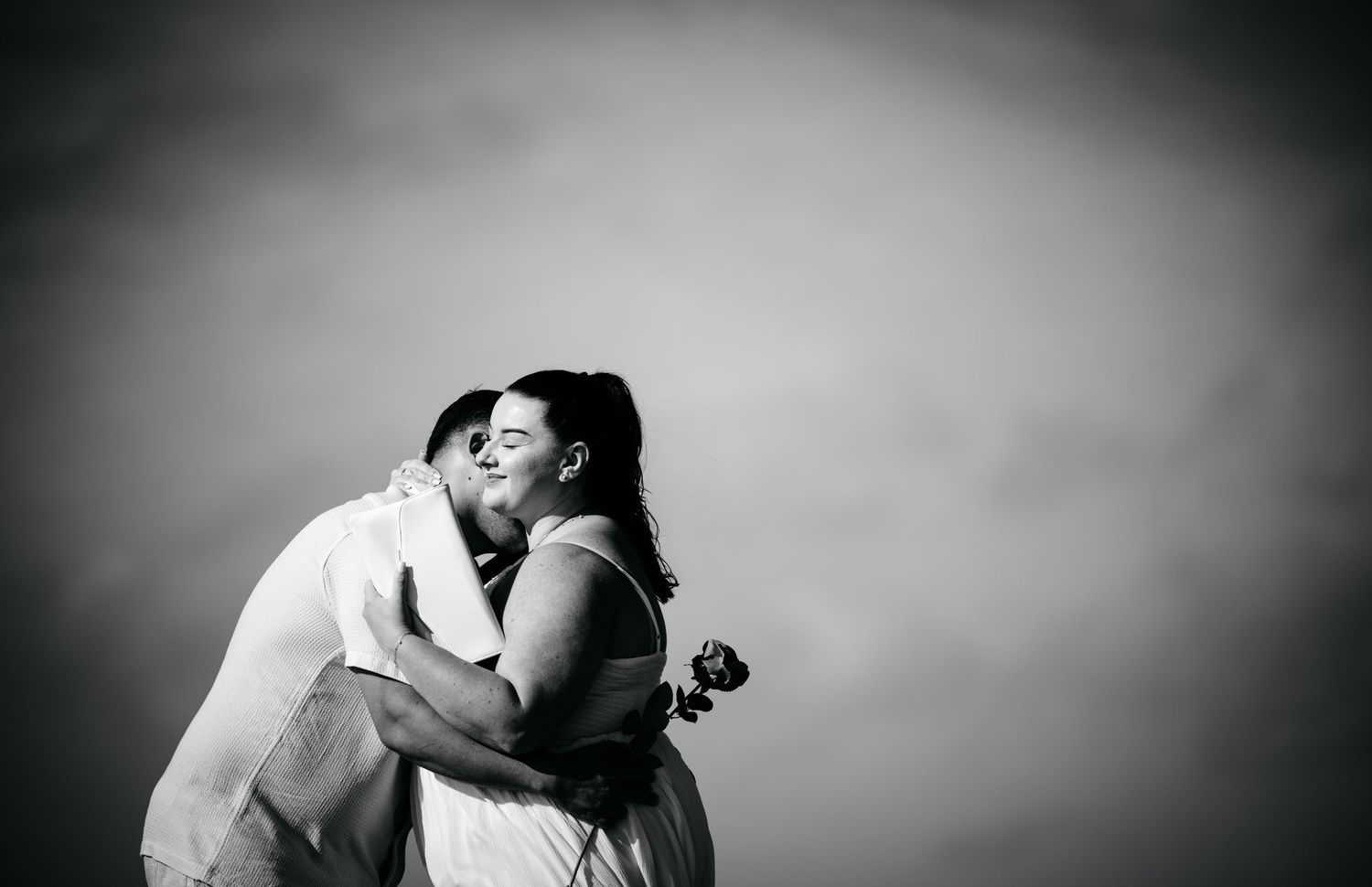 Black and white photo of couple embracing and kissing against dramatic cloudy sky backdrop.