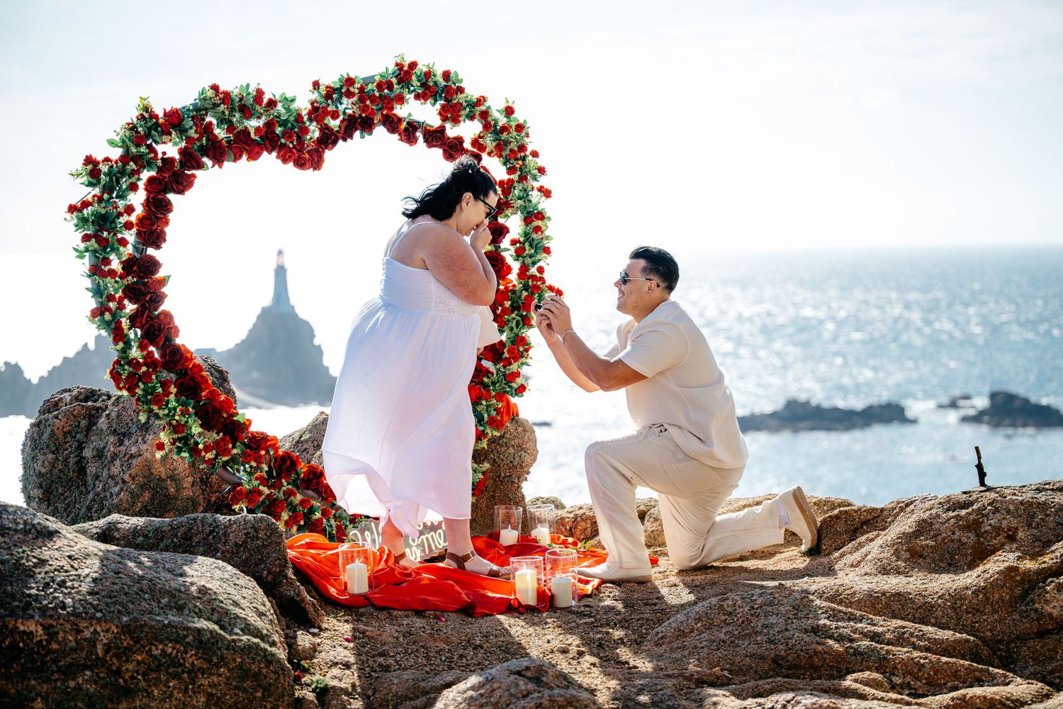 Man proposes to woman beside heart-shaped floral arch on rocky coastal clifftop at sunset.