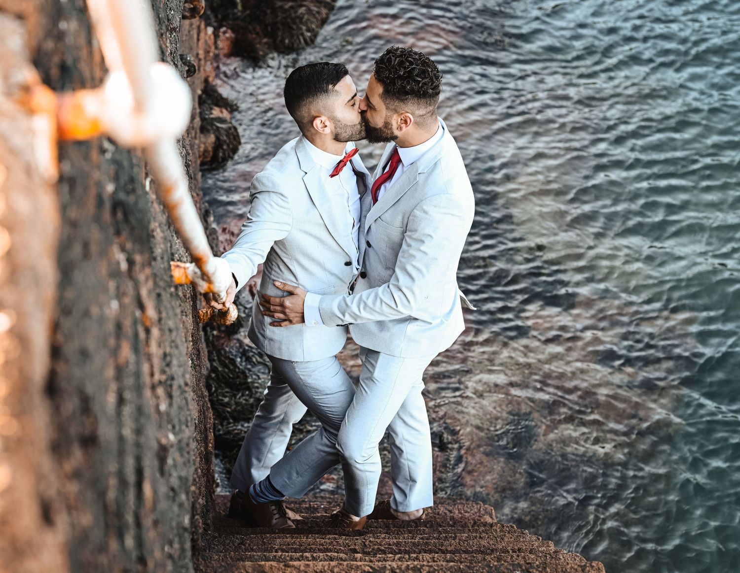 Two grooms in white suits share a kiss on stone steps beside the ocean.