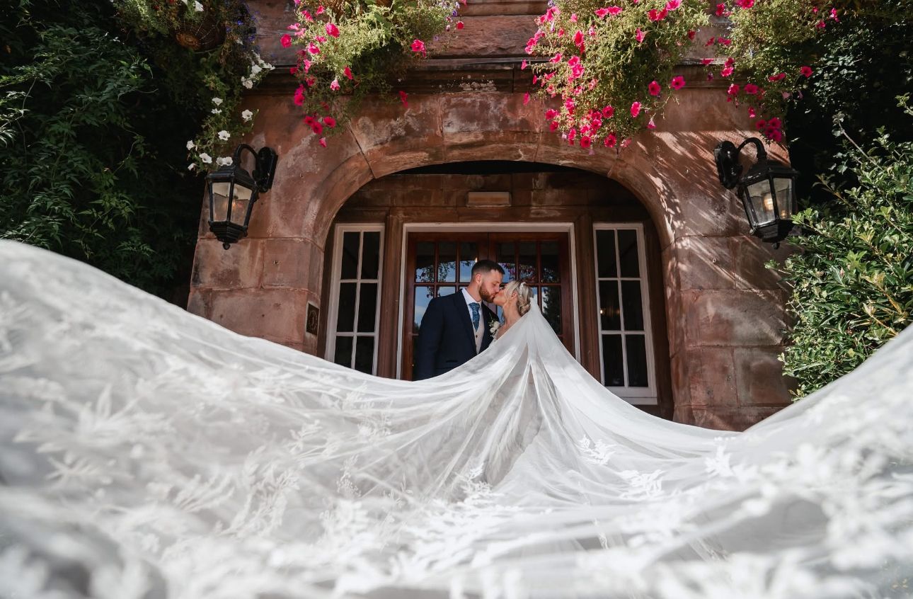 Couple shares romantic kiss under floral archway with bride's lace veil dramatically spread wide.