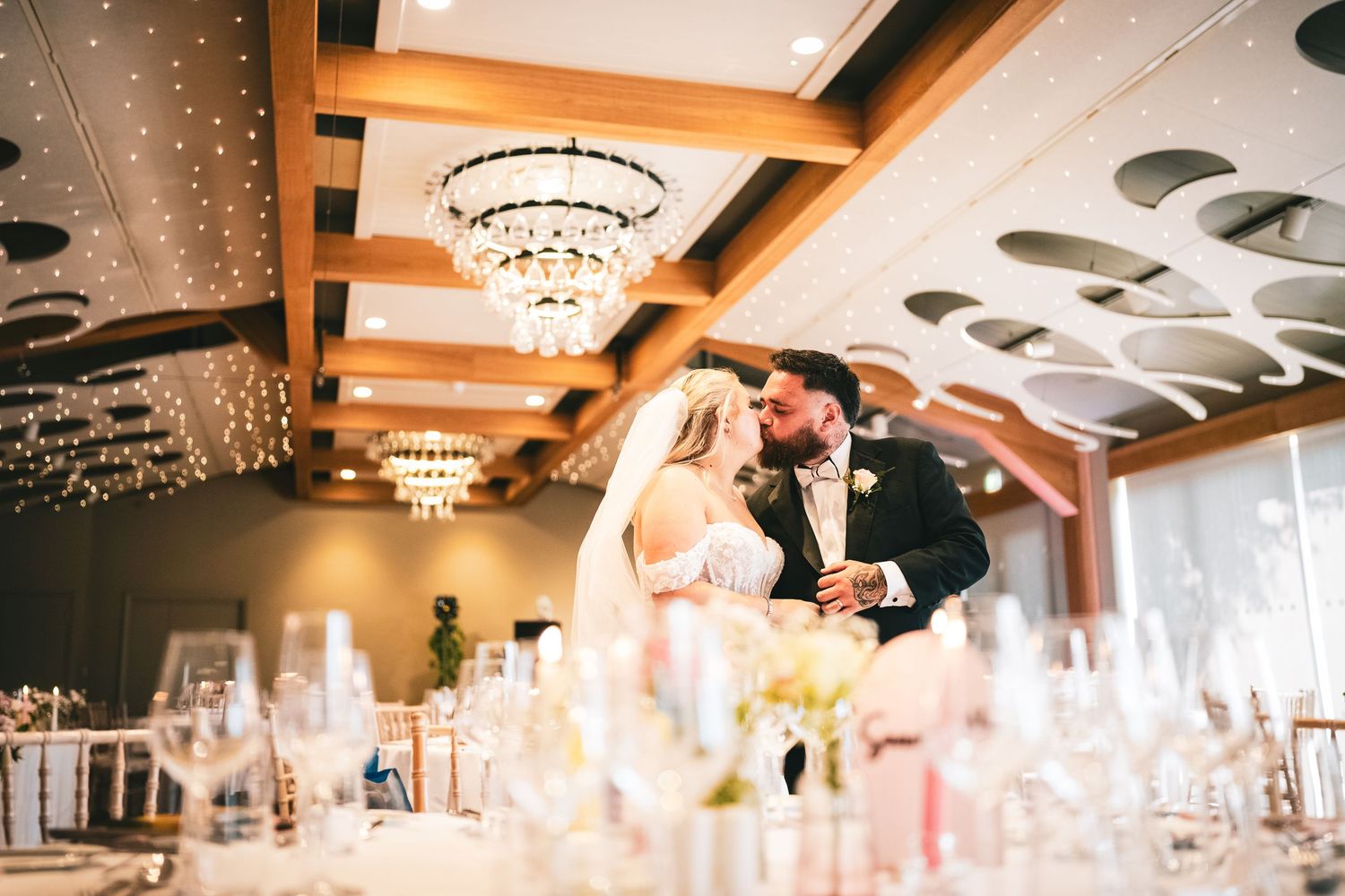 Bride and groom share romantic kiss in elegant chandelier-lit wedding reception ballroom.