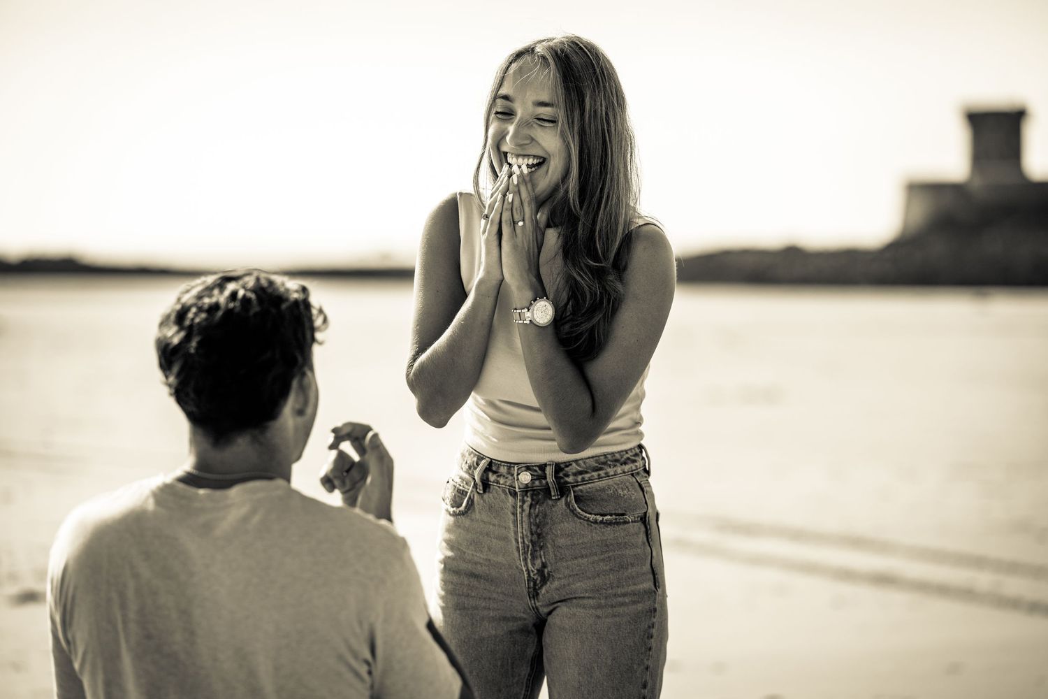 Man proposing to surprised woman on beach, she covers mouth with hands in joyful shock.