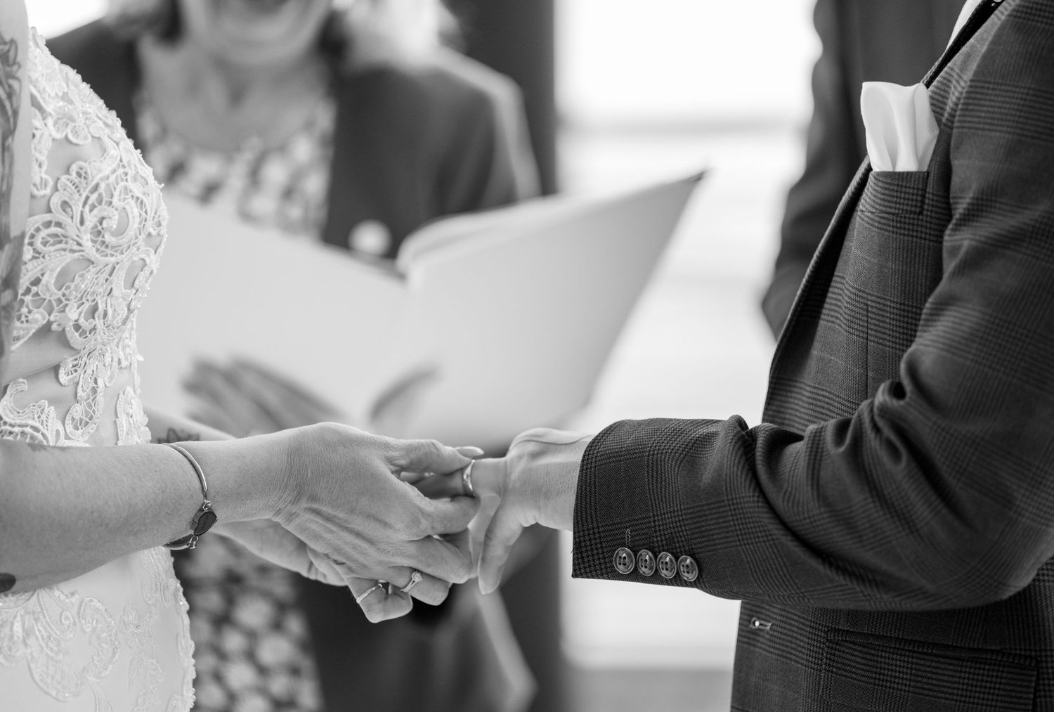 Black and white close-up of couple exchanging rings during wedding ceremony with officiant holding book.