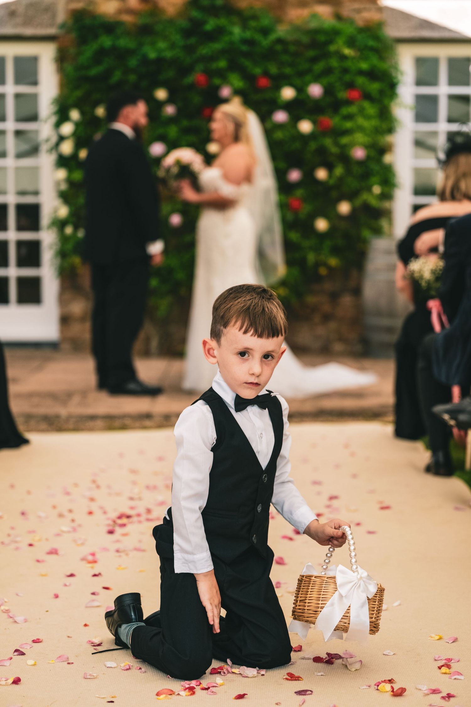 Young ring bearer in tuxedo kneels on petal-strewn aisle while bride and groom exchange vows.