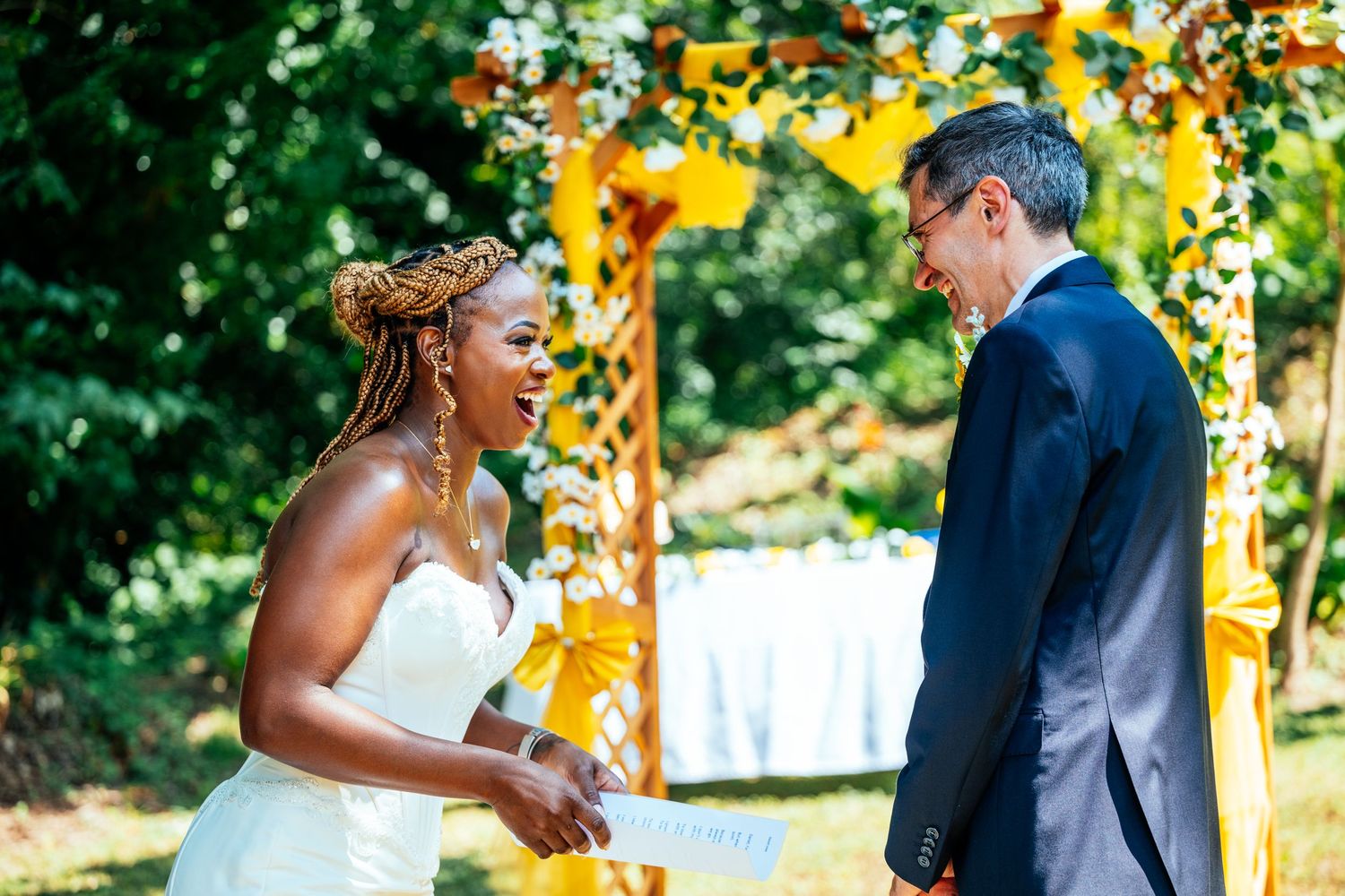 A bride in a white strapless gown laughs joyfully with her groom during an outdoor ceremony with yellow floral decor.