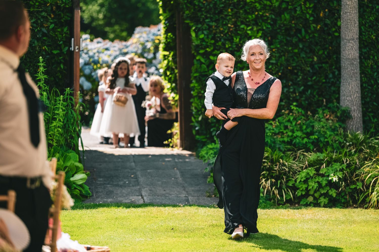 A woman in black carries a baby boy as flower girls in white dresses walk along a garden path at a wedding.
