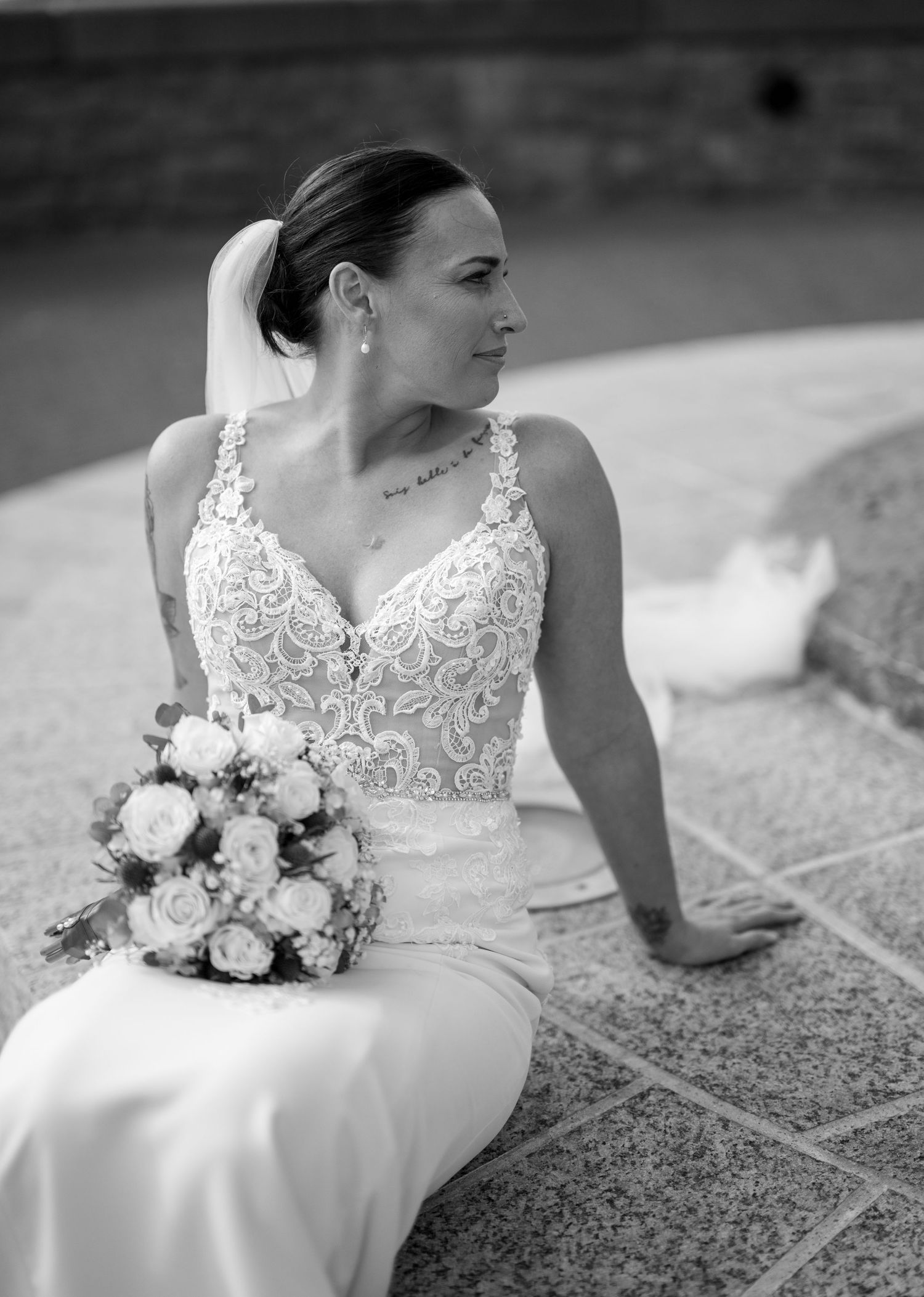 Black and white portrait of bride in lace gown sitting with bouquet, showing shoulder tattoo.