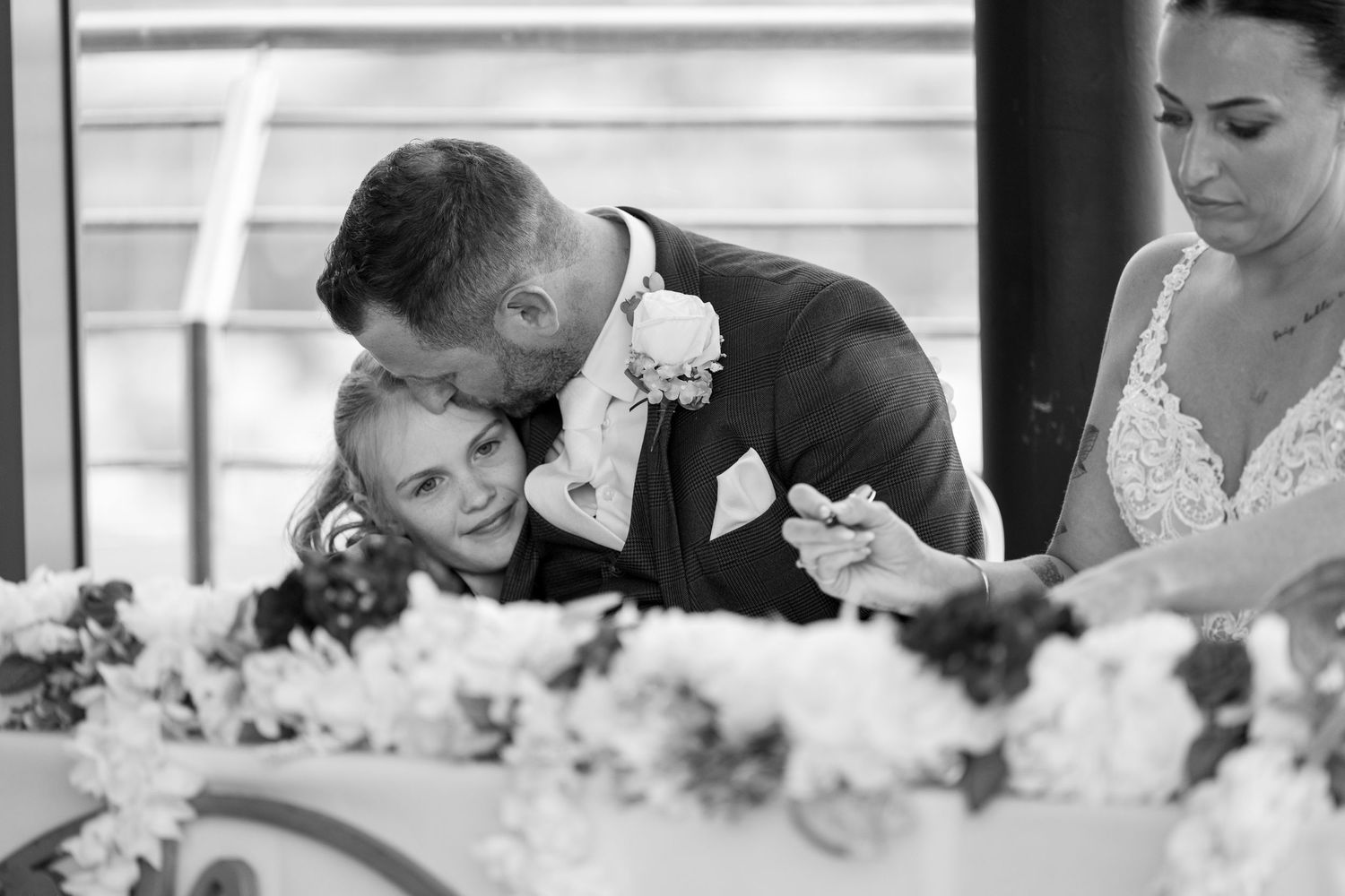 Black and white photo of groom embracing young girl at wedding ceremony table with bride nearby.