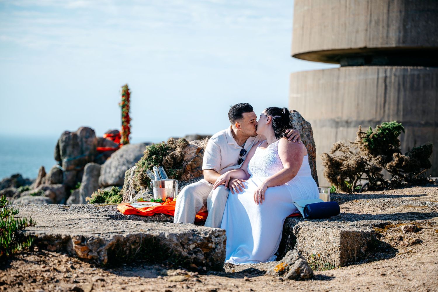 Couple shares romantic kiss while seated on coastal rocks with ocean view behind them.