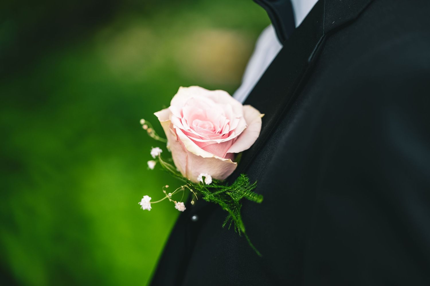 A delicate pink rose boutonniere with baby's breath and greenery pinned to a black tuxedo lapel.