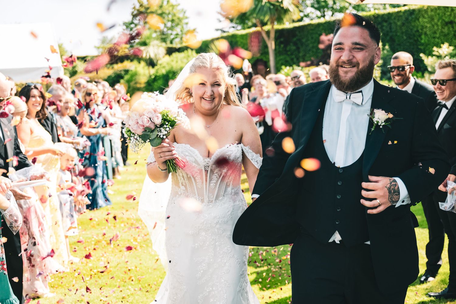 Joyful newlywed couple walks down aisle as guests toss colorful flower petals in celebration.