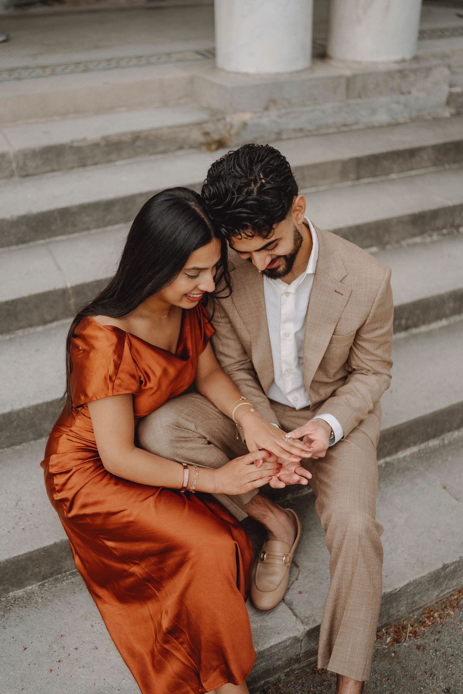 Couple sitting on stone steps holding hands and looking down at an engagement ring together.