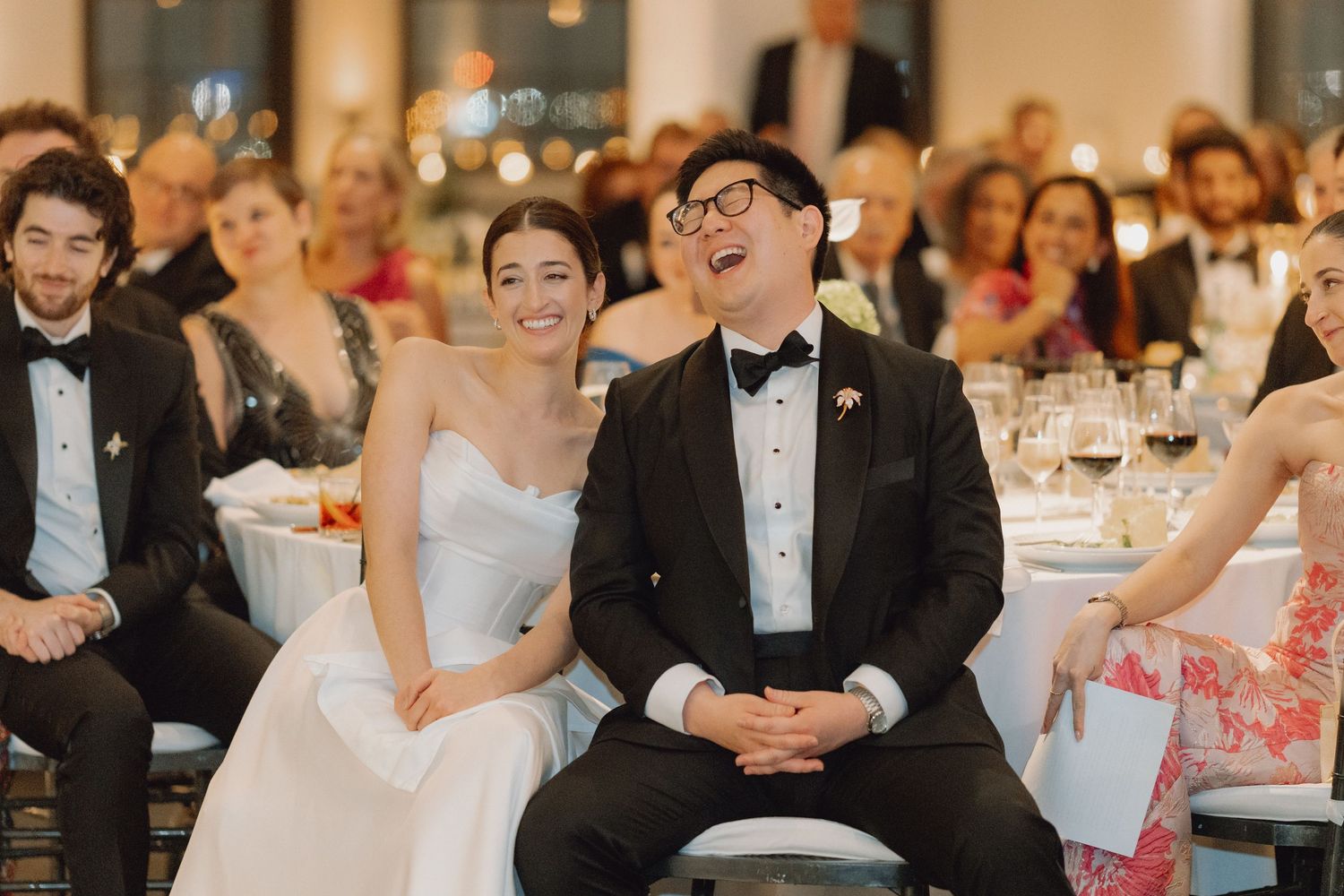 Bride and groom laugh joyfully while seated at elegant wedding reception surrounded by guests.