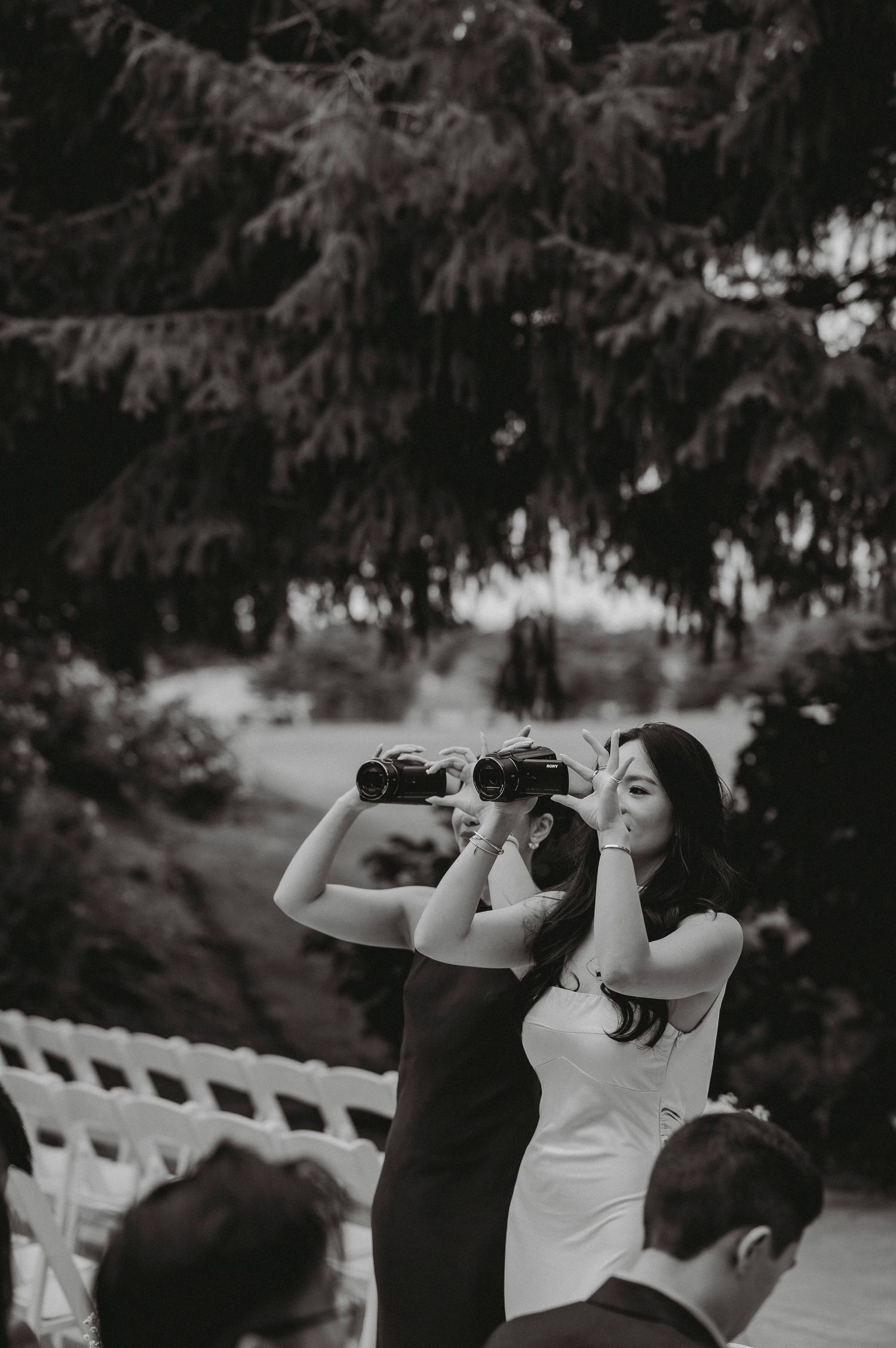 Two women use binoculars at outdoor wedding ceremony in playful black and white candid photo.