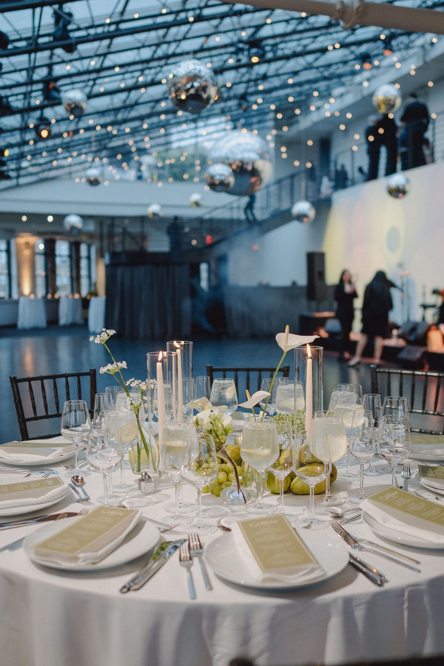 Elegant wedding reception table with white linens, candles, and greenery centerpieces under disco balls in an industrial venue.