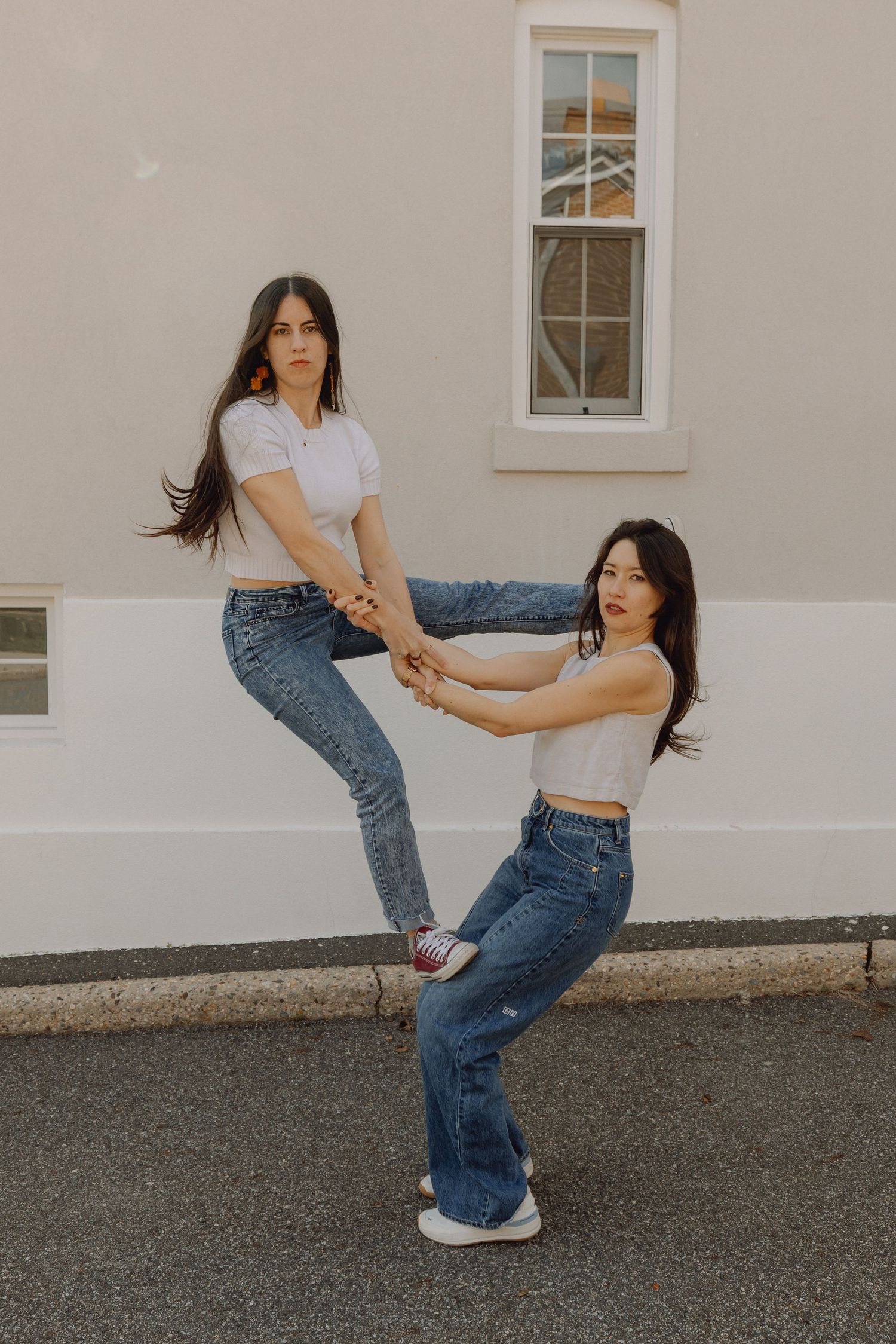 Two young women in white tops and jeans hold hands while posing outside a building.