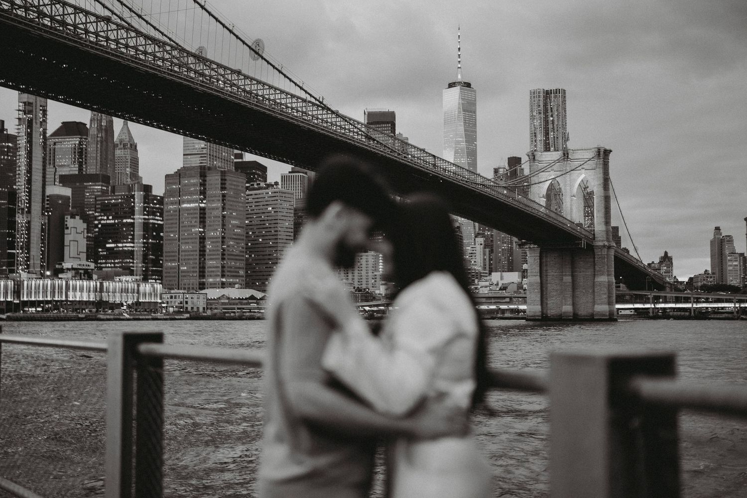 Black and white photo of a couple embracing near the Brooklyn Bridge with the NYC skyline in the background.