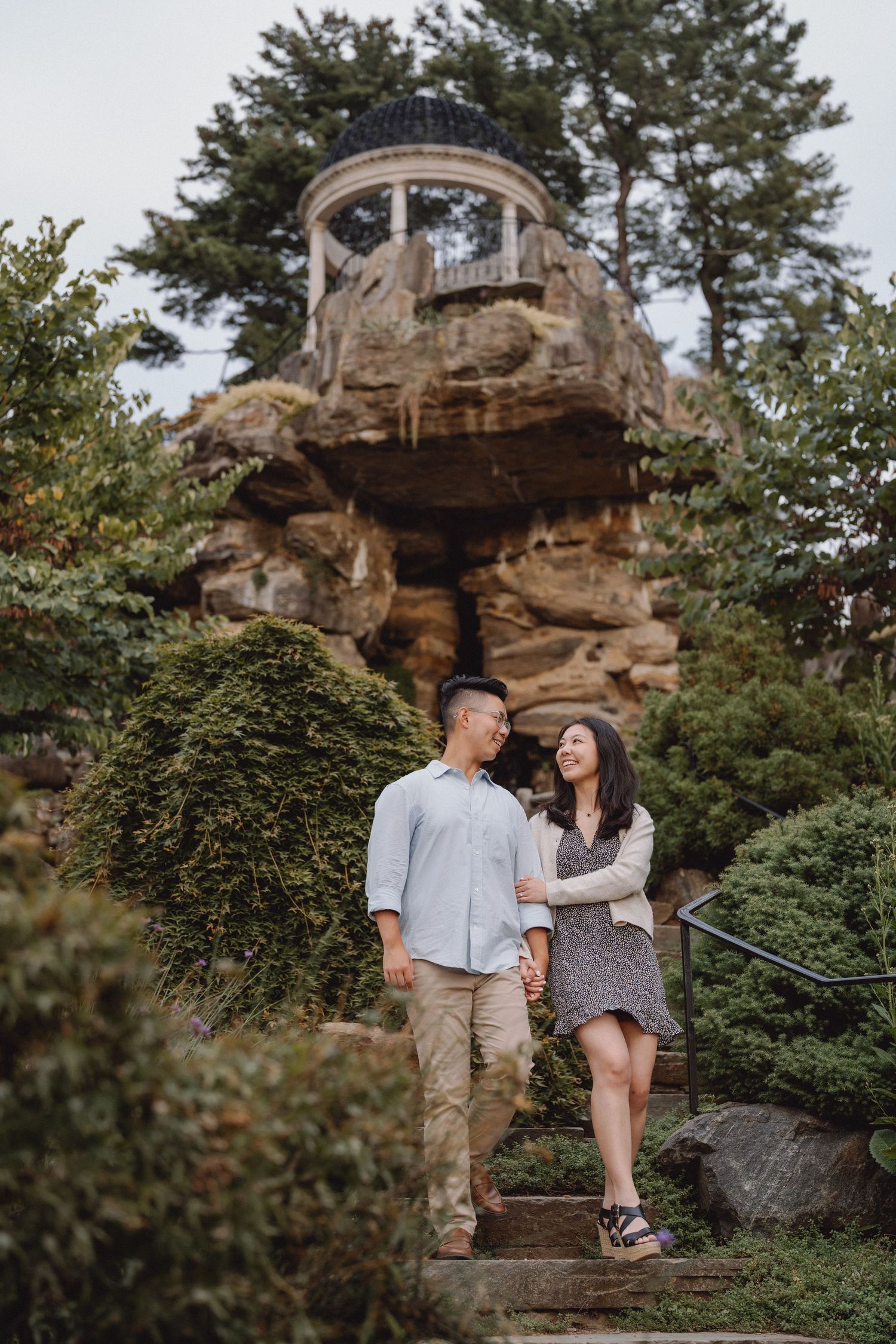 Smiling couple holding hands and walking down stone steps in a lush garden with a rocky hillside gazebo behind them.