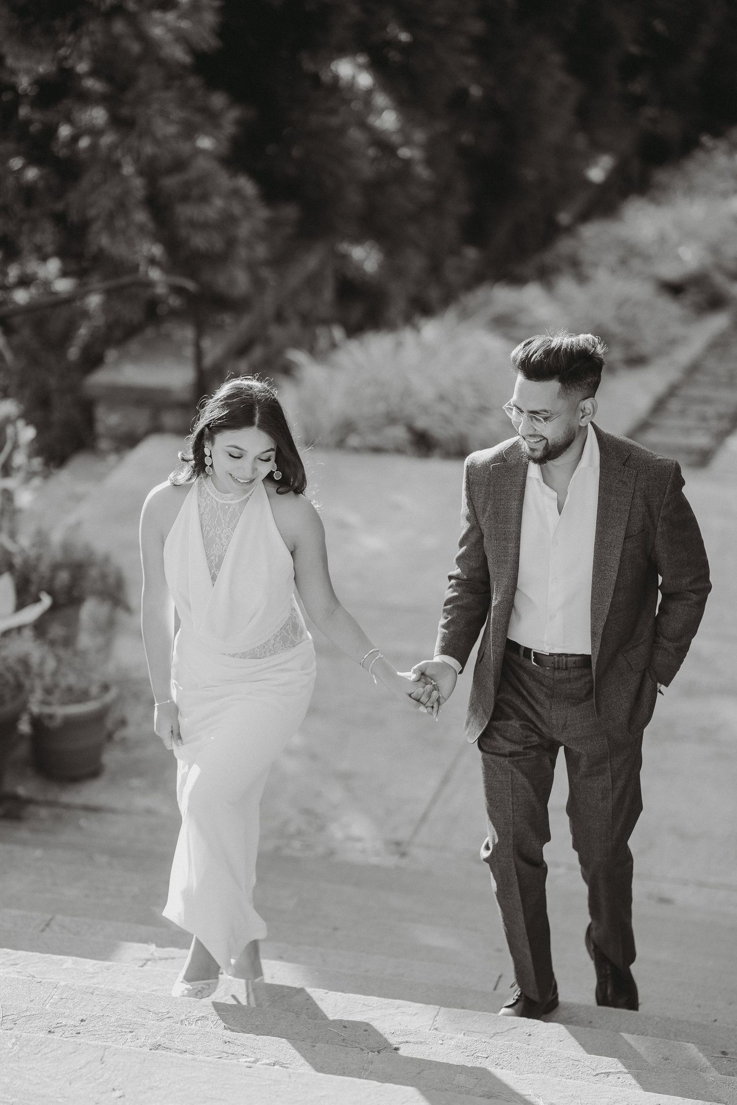 Black and white photo of a smiling couple holding hands and walking up outdoor stone steps, she in a white halter dress, he in a suit.