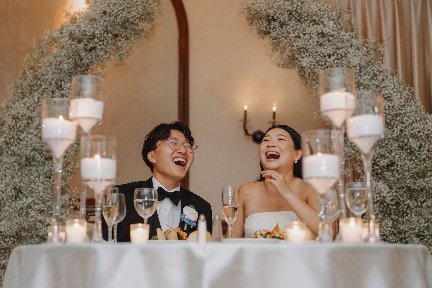 Bride and groom laugh joyfully at sweetheart table decorated with baby's breath floral arch and candles.
