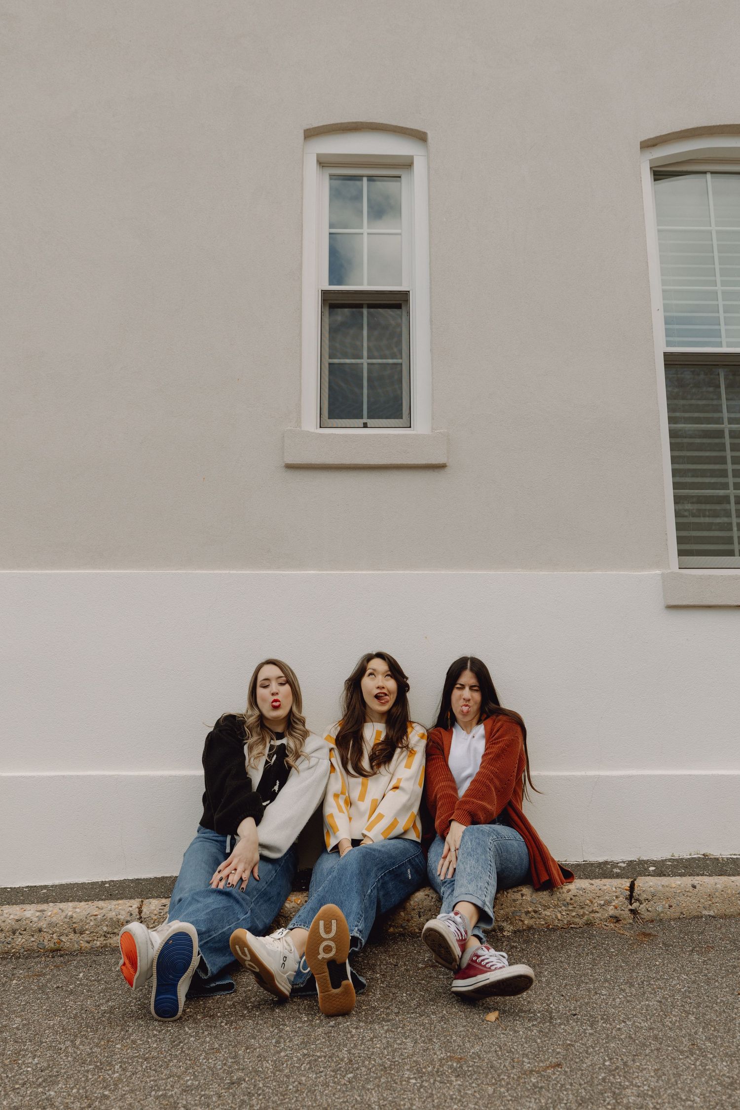 Three young women sitting against a building wall in jeans and colorful sneakers making fun expressions.