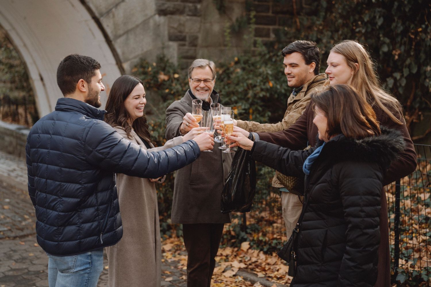 Group of six friends in winter coats toasting with glasses of champagne outdoors near a stone archway on a leaf-covered path.