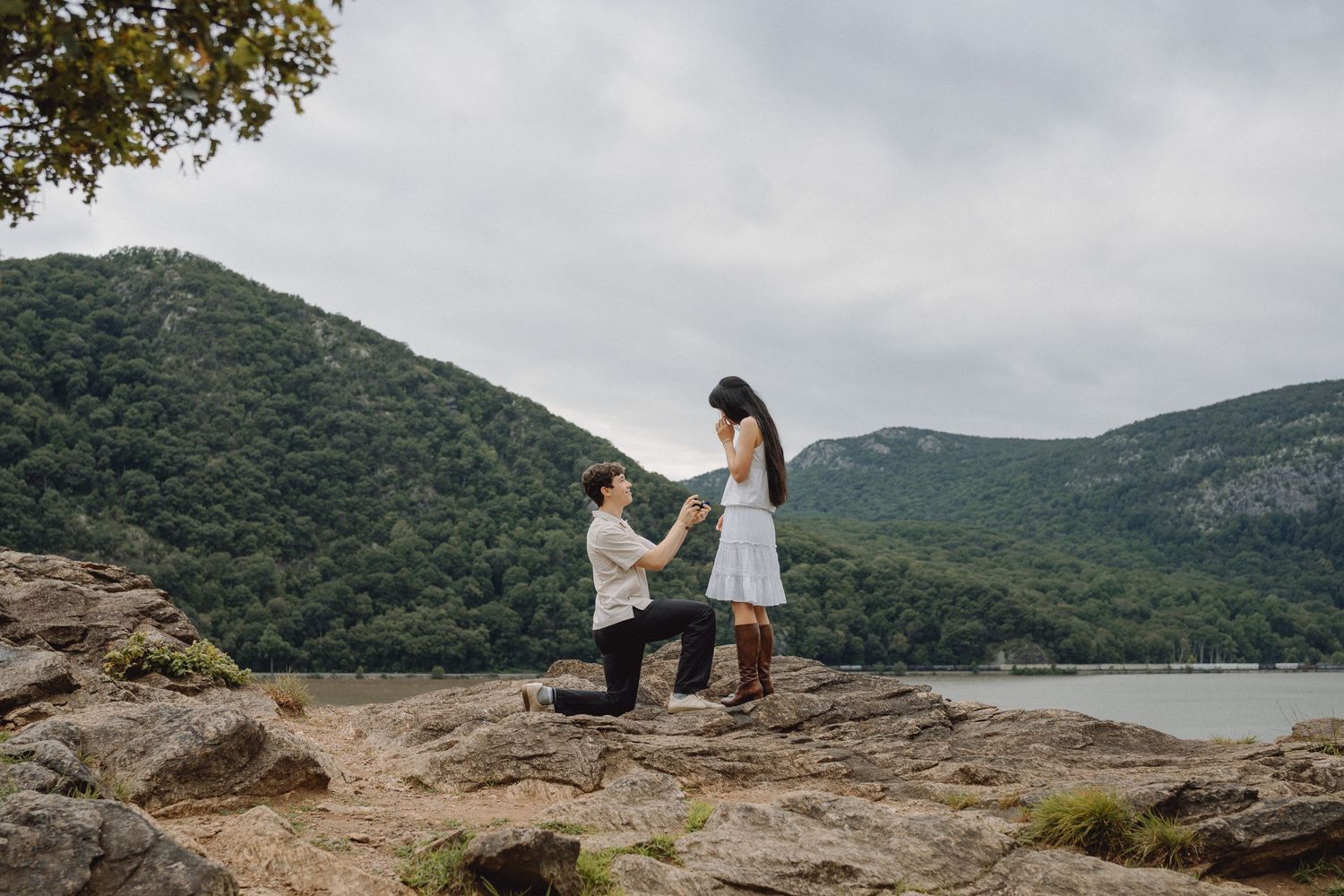 Man kneels on a rocky overlook proposing to a woman in a white dress with forested mountains and a lake in the background.