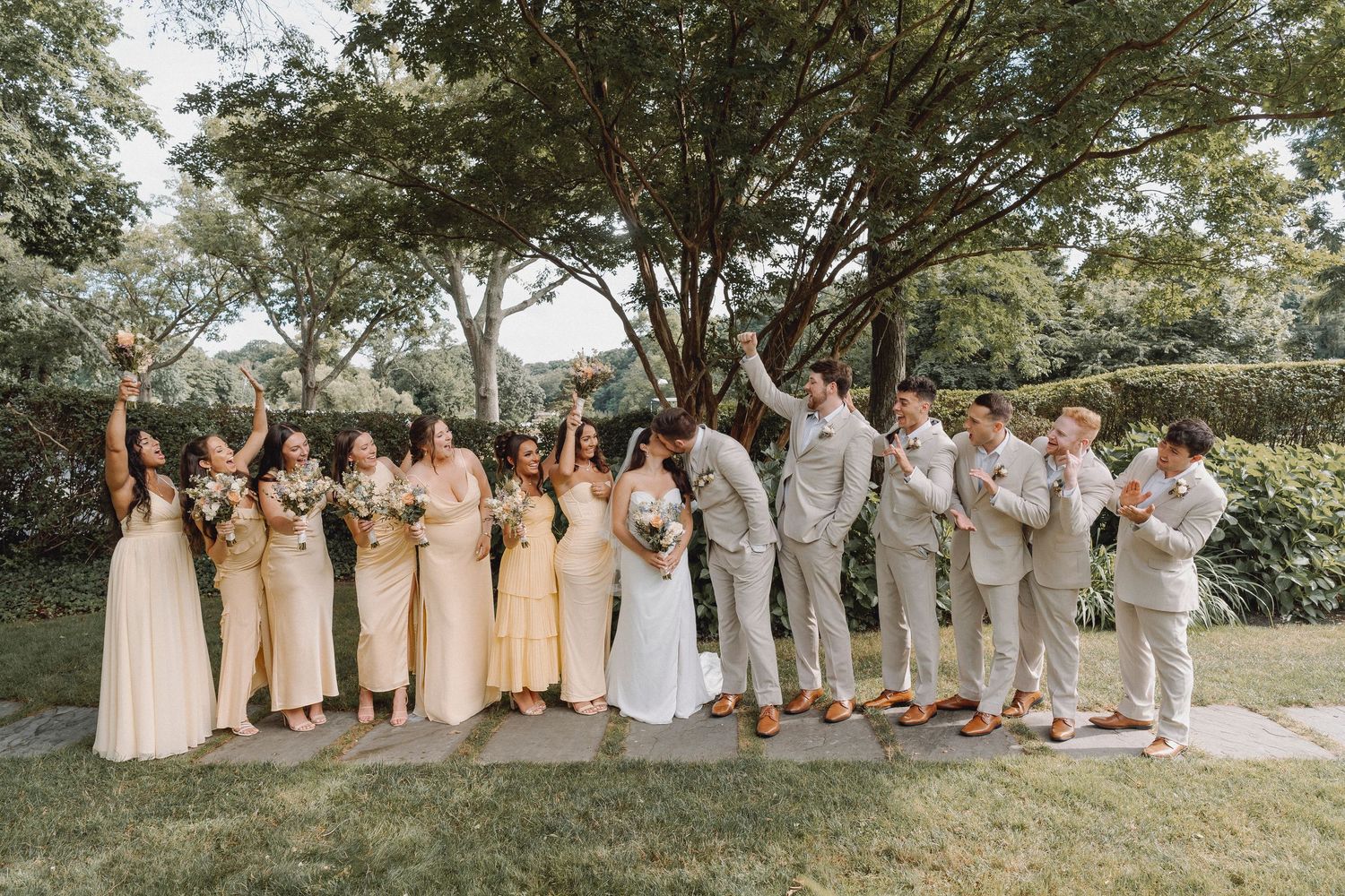 Wedding party poses outdoors under trees, bridesmaids in yellow dresses and groomsmen in tan suits.