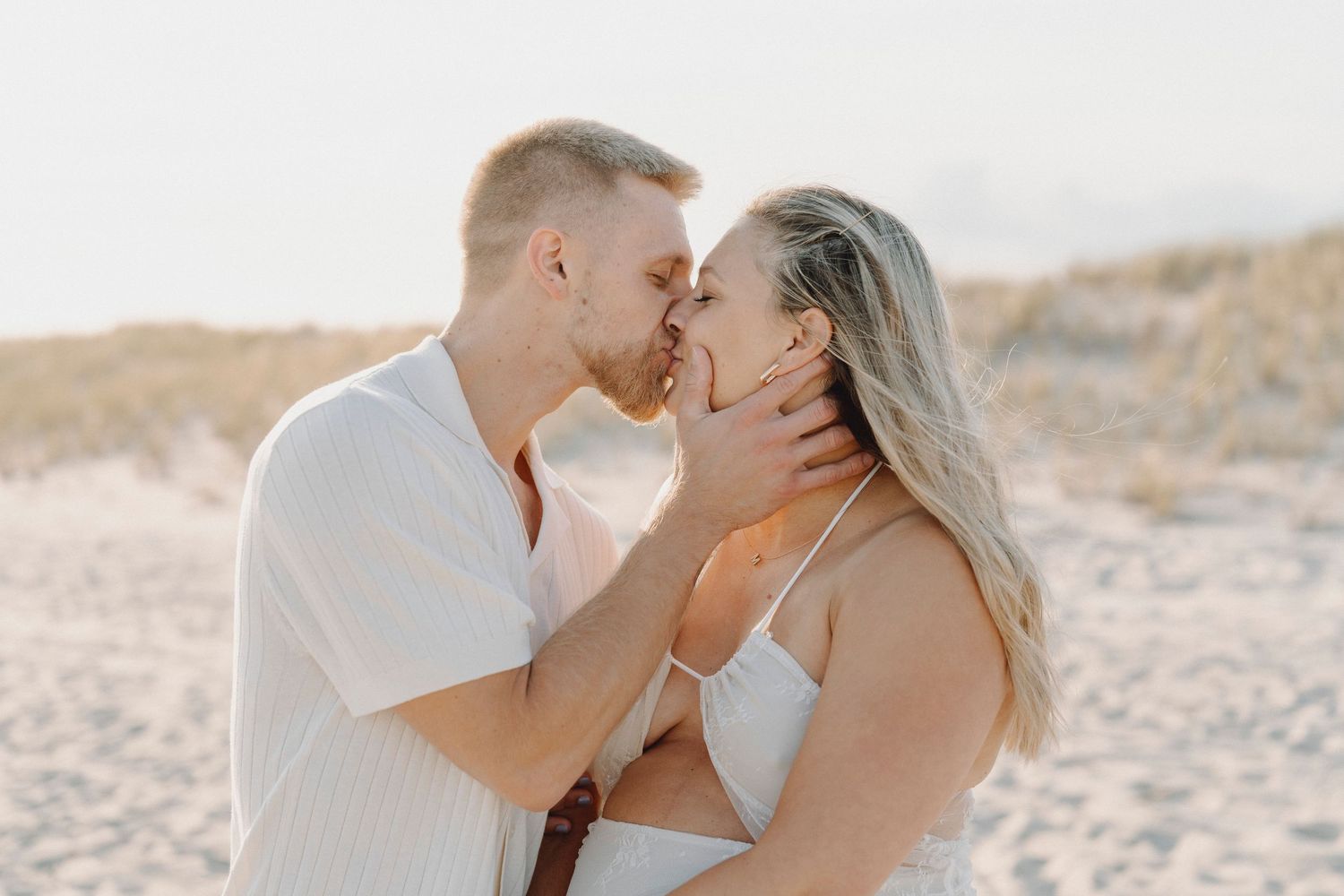 Couple sharing romantic kiss on sandy beach with grass dunes in background.