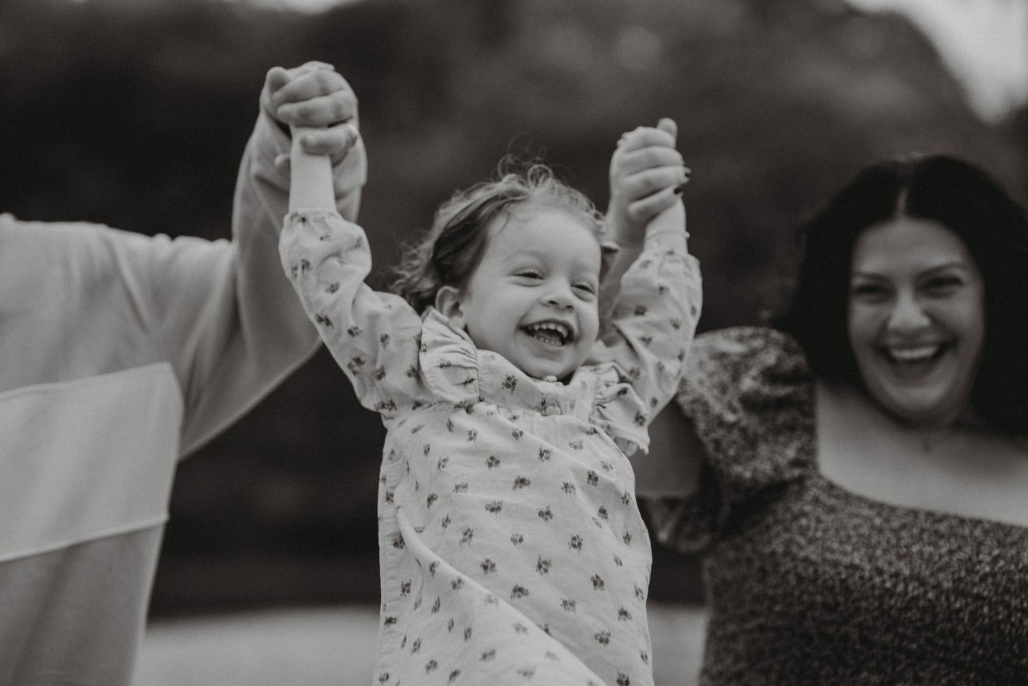 Black and white photo of a laughing toddler girl with arms raised held by her smiling parents outdoors.