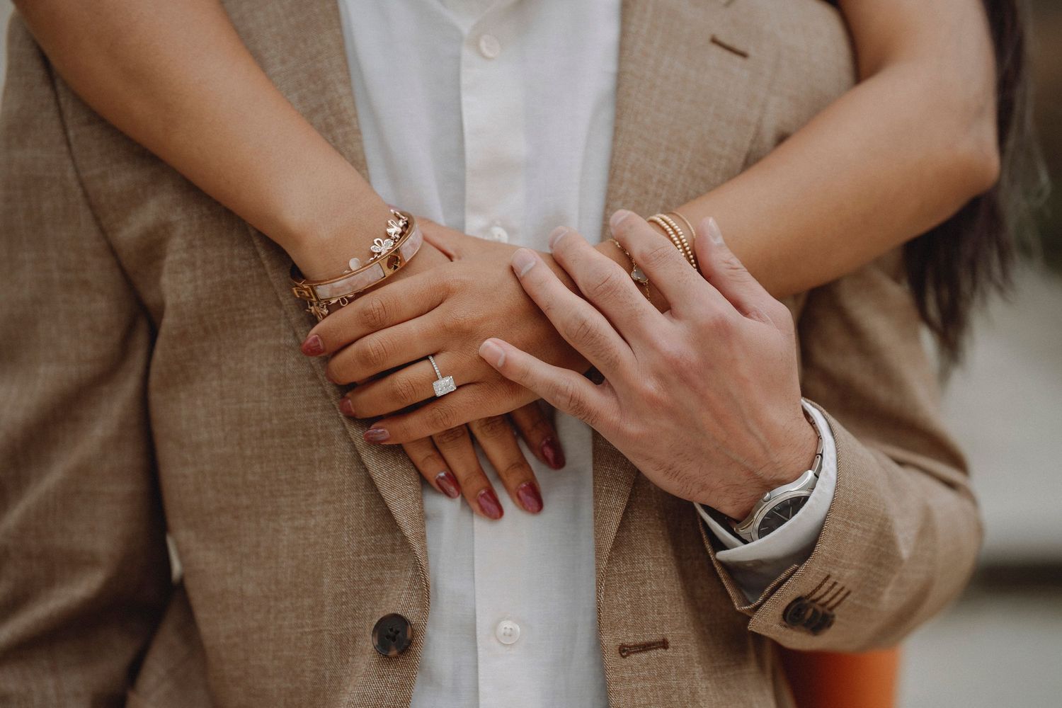 Close-up of couples hands intertwined, wearing gold bracelets and rings, man in beige suit, woman in rust dress.