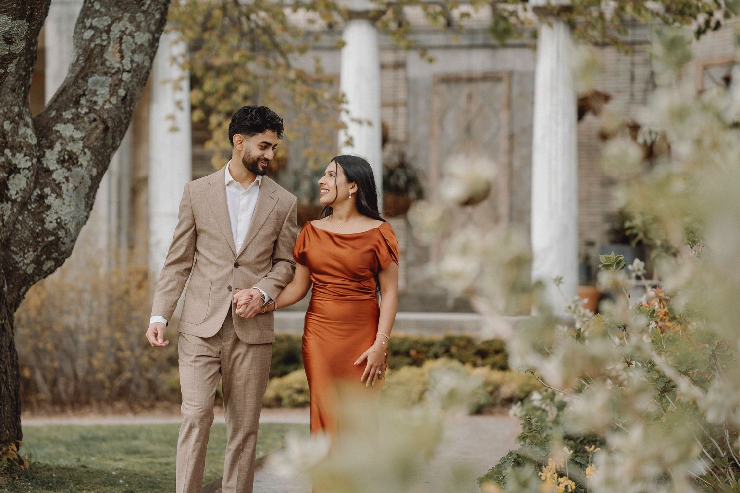 Couple in rust dress and beige suit walking hand in hand among blooming trees by columns.