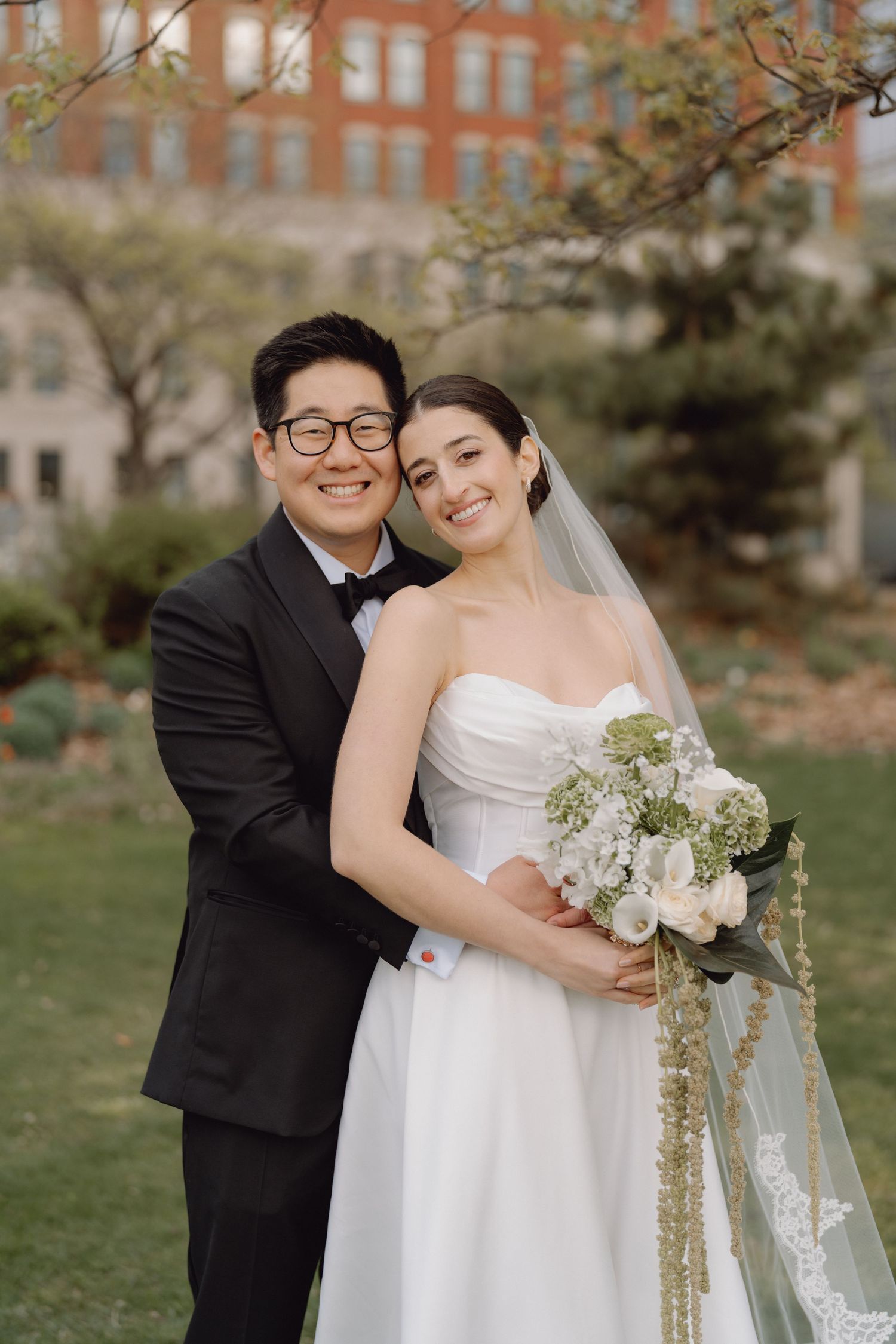 Smiling bride and groom pose outdoors in garden setting, bride holding white and green bouquet.
