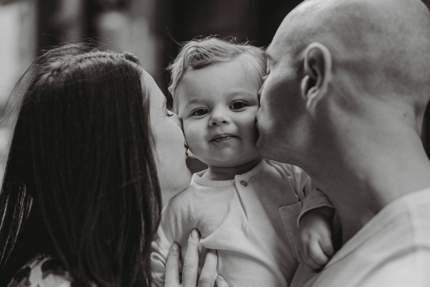 Parents kissing their smiling baby on the cheeks in a tender black and white portrait.