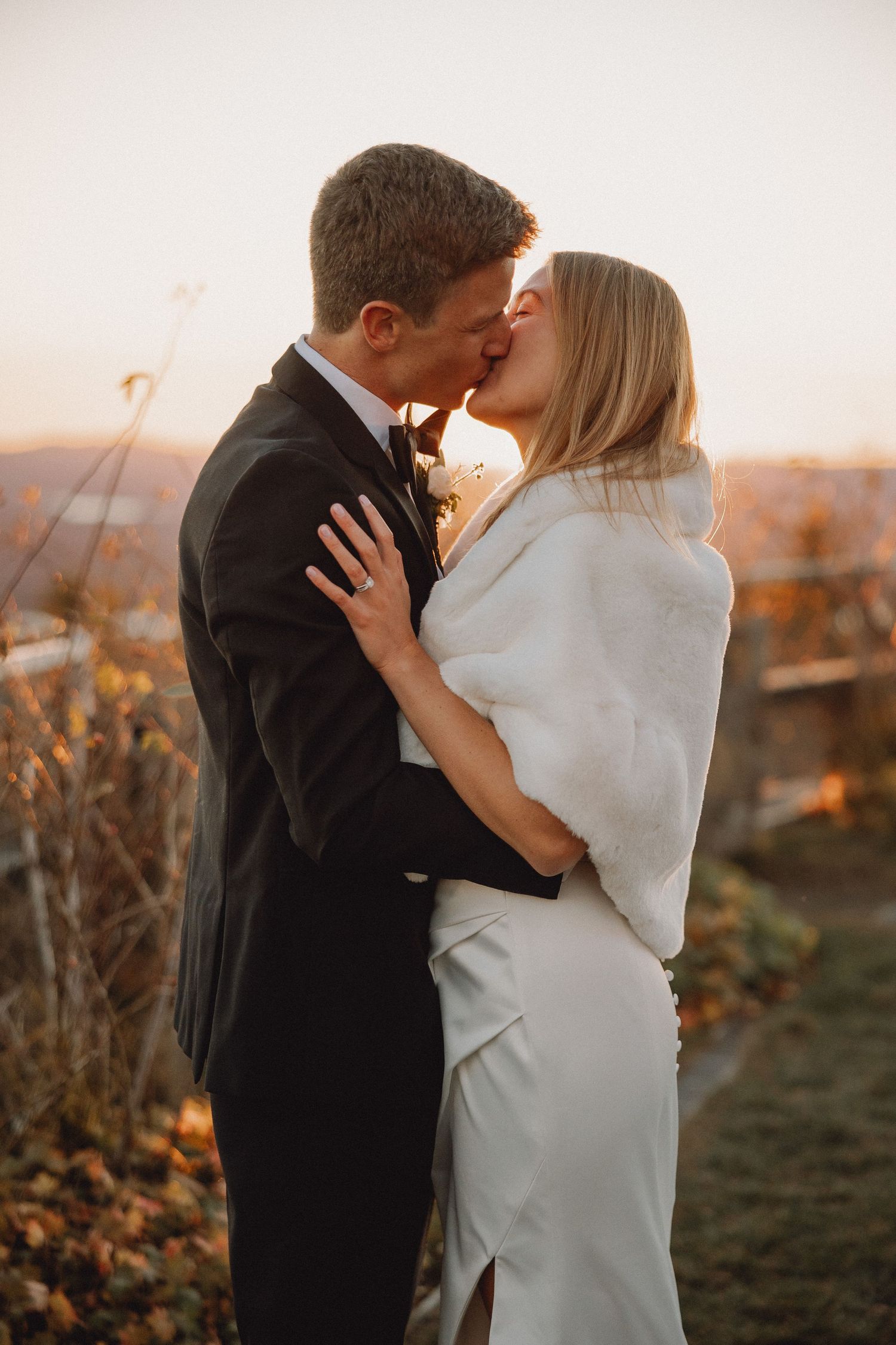 Newlyweds kissing at golden hour sunset, bride in white fur stole and fitted gown, groom in classic black suit outdoors.