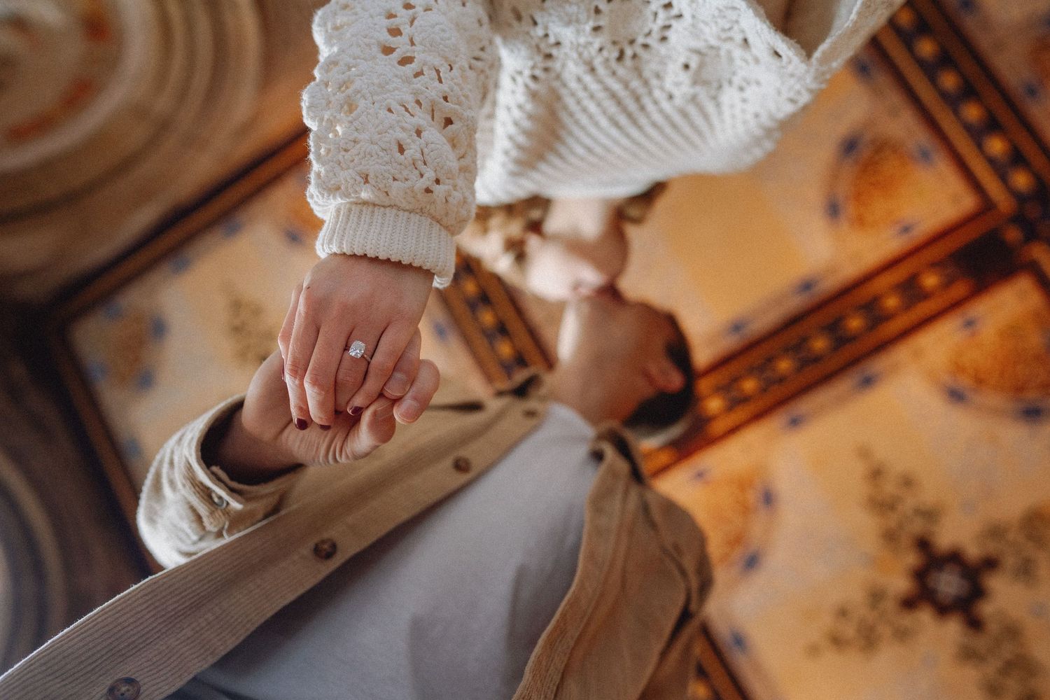 Close-up of a woman's hand with an engagement ring holding her partner's hand on a ornate surface.