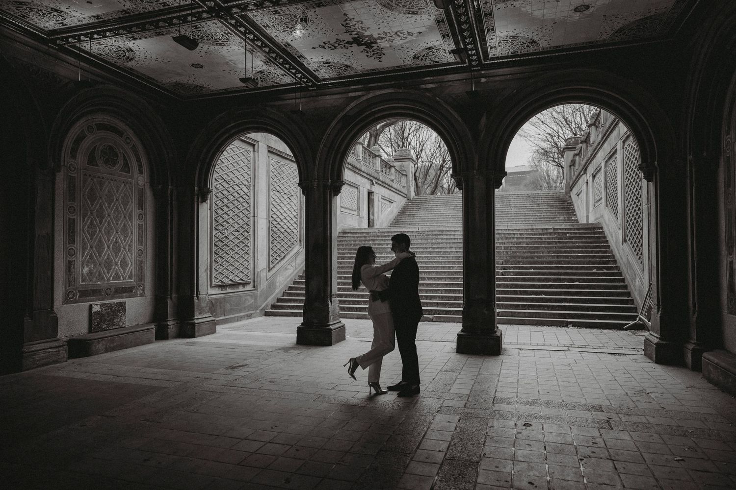 Black and white photo of a couple embracing under stone arches at Bethesda Terrace in Central Park.