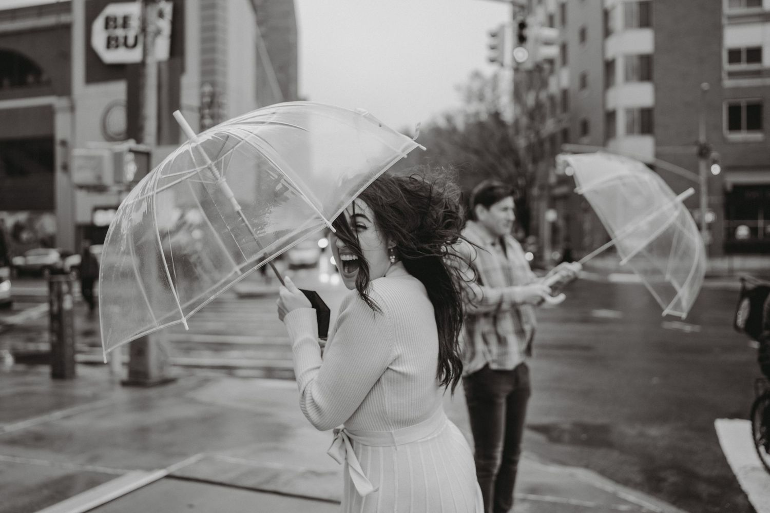 Black and white photo of woman with clear umbrella on rainy urban street.