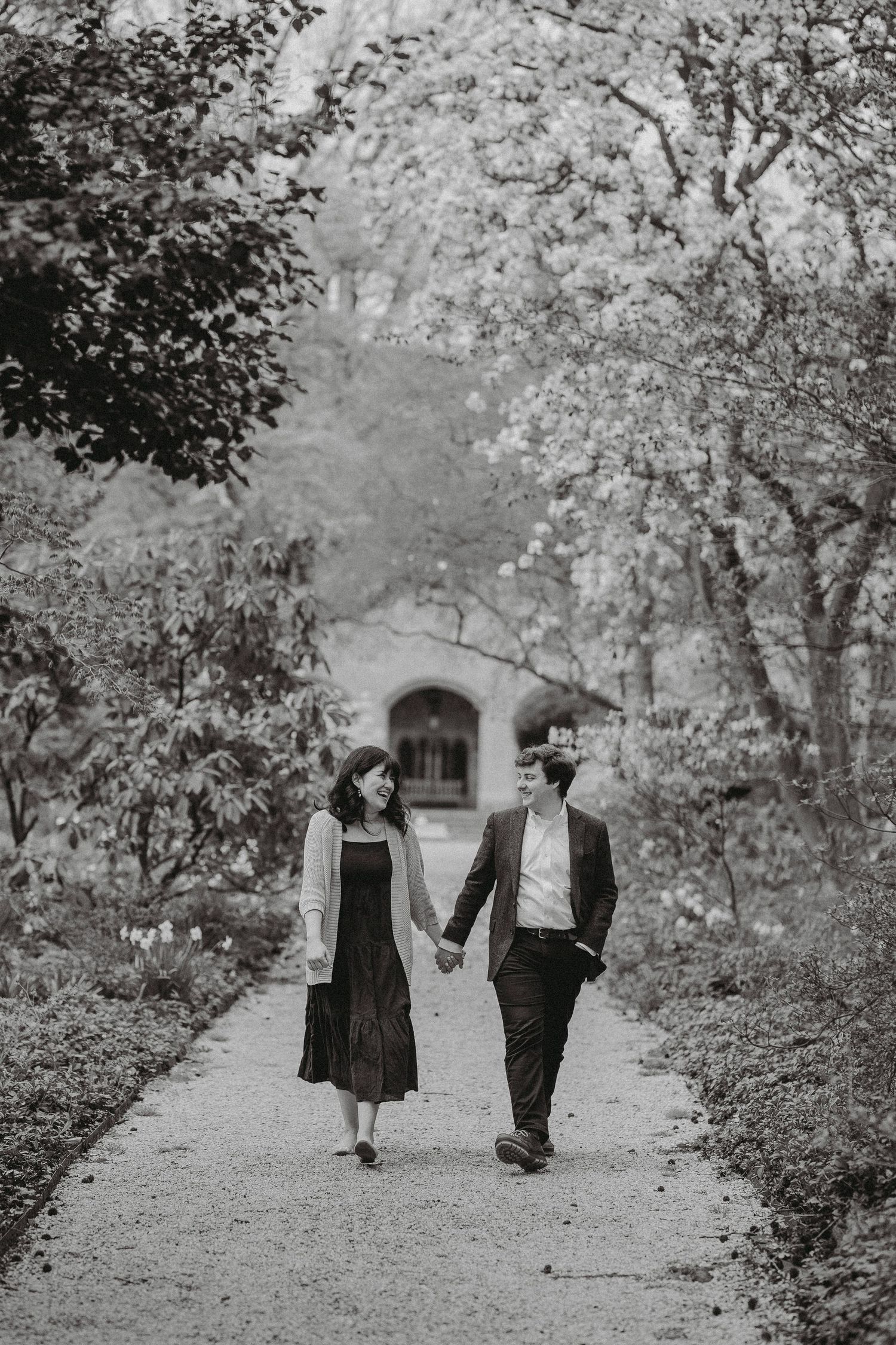 Black and white photo of an engaged couple walking hand in hand along a garden path.