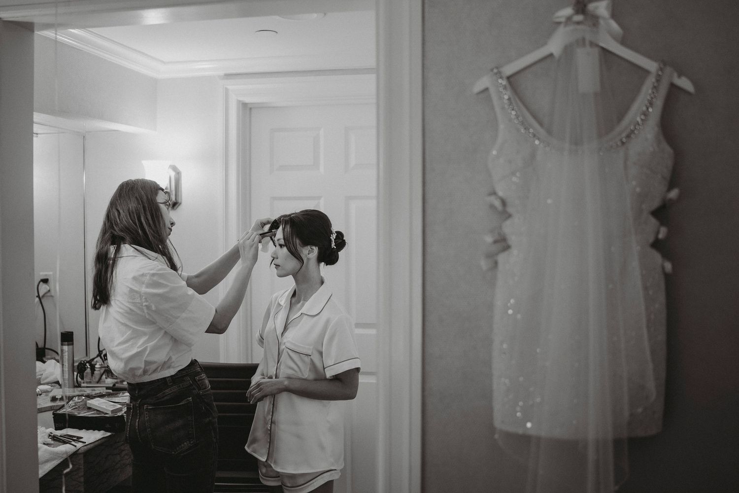 Black and white photo of bride getting hair done with wedding dress hanging nearby.